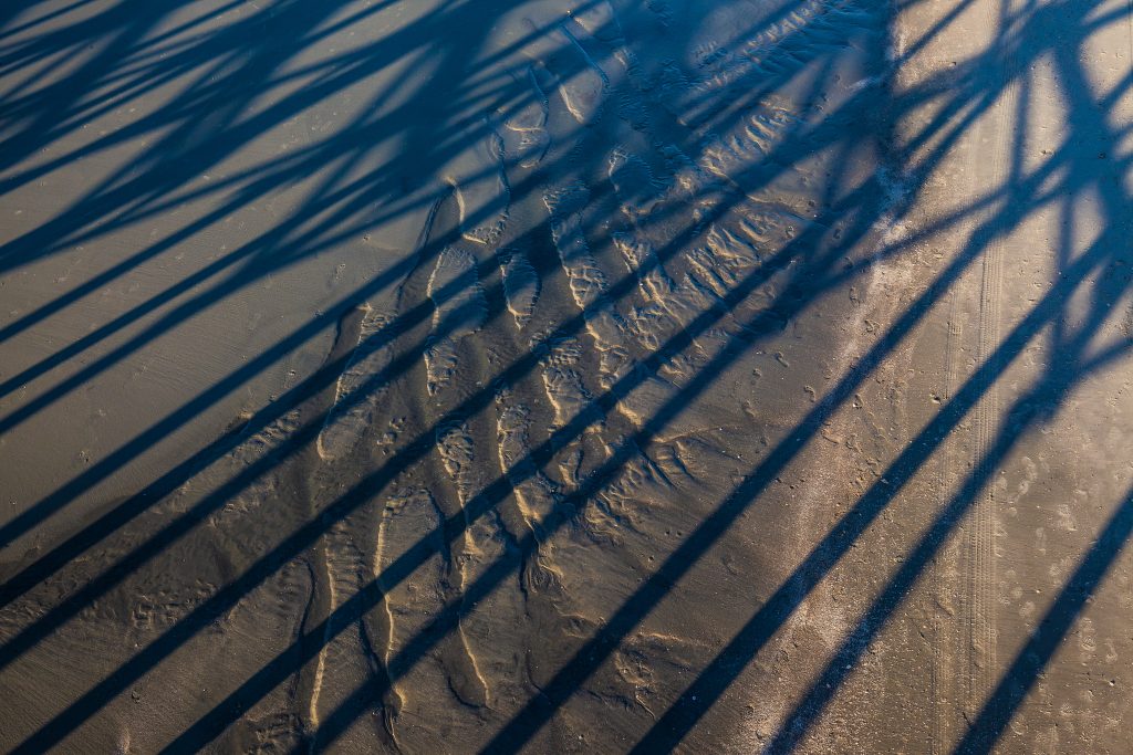 Abstract image examining the play of shadows from a pier upon the erosional patterns in the sand.