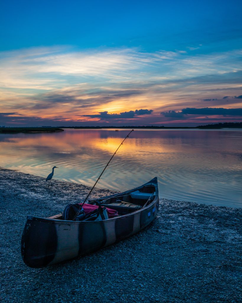 Image is of a fishing canoe on the water's edge of a salt marsh at sunrise as an egret poses.